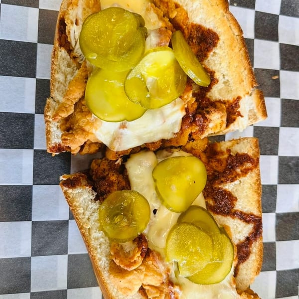 Close-up of a fried chicken sandwich with melted cheese and pickles on a checkered paper.
