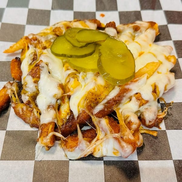Cheesy fries topped with melted white cheese and sliced green pickles on a checkered paper.