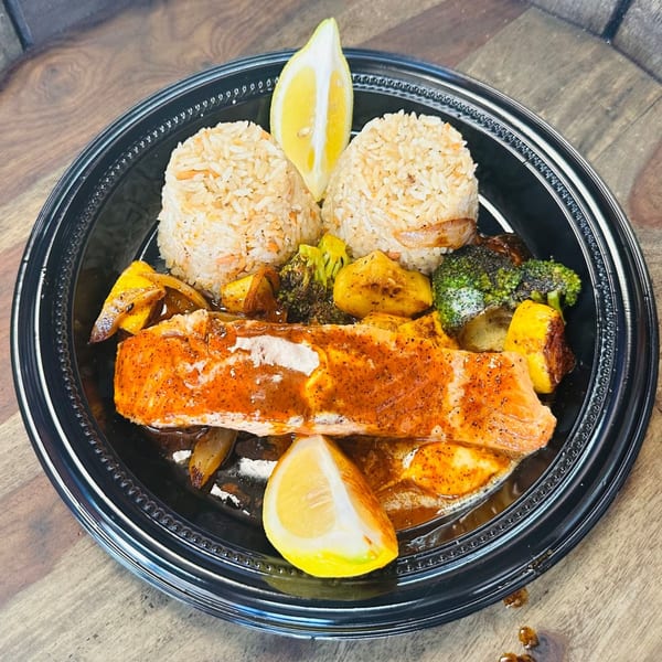 Salmon fillet with rice, roasted broccoli, potatoes, and lemon wedges in a black takeout container.